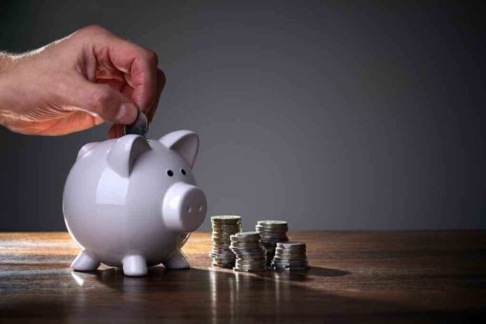 A hand inserting a coin into a white piggy bank on a wooden table with stacks of coins nearby