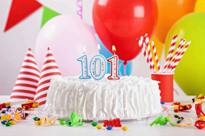 A decorated birthday cake with the number 101 and colorful balloons in the background