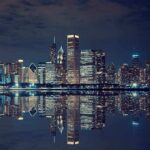 Illinois Governor BLOCKS Trump’s Chicago Crime Crackdown Nighttime view of a city skyline with reflections on water