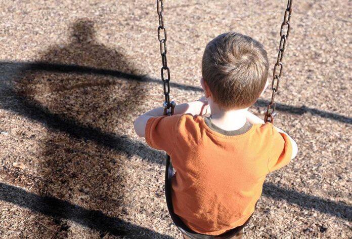 A young boy sitting on a swing at a playground with his shadow cast on the ground