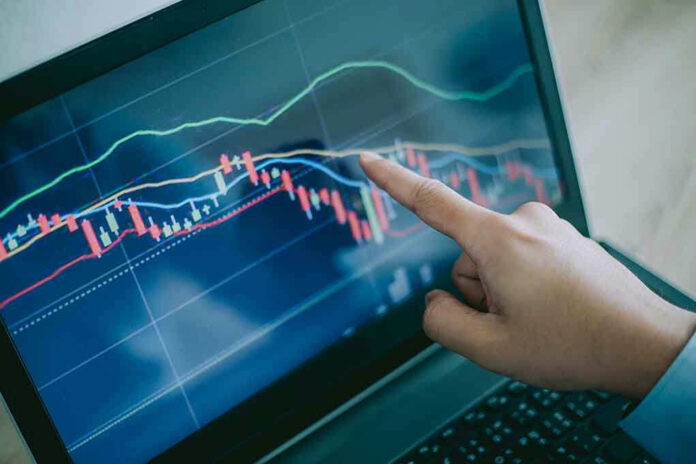 Hand pointing at stock market graph on laptop screen.