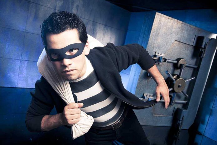 A masked thief in a striped shirt holding a bag while emerging from a bank vault