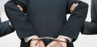 Person handcuffed in suit, escorted by two others.