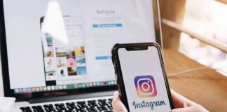 Person holding a smartphone displaying the Instagram logo with a laptop in the background