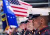 U.S. Marines in uniform standing in formation with flags in the background