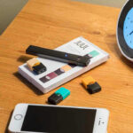Vape pen, smartphone, clock on wooden table.