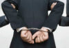 Person handcuffed in suit, escorted by two others.