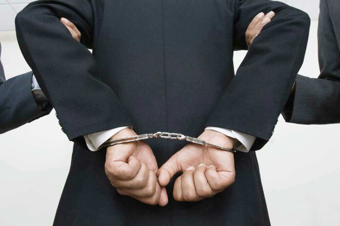 Person handcuffed in suit, escorted by two others.