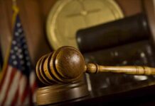 A wooden gavel resting on a desk in a courtroom with an American flag in the background