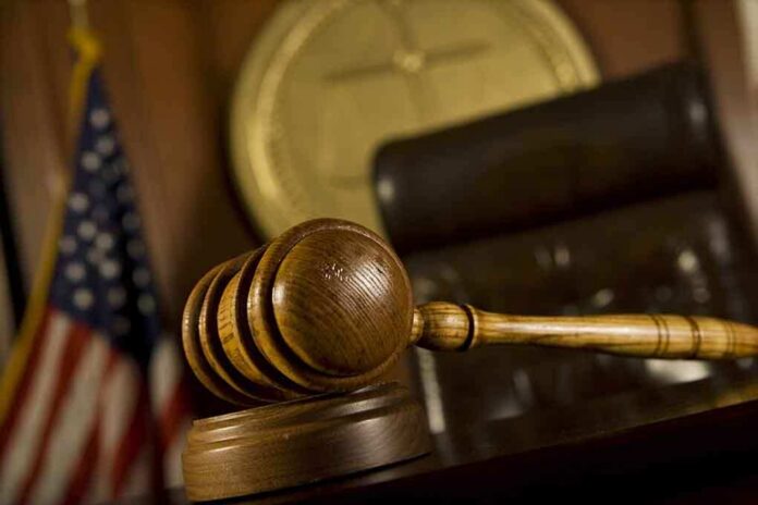 A wooden gavel resting on a desk in a courtroom with an American flag in the background