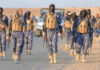 Camouflaged soldiers wearing masks and vests walk in desert.