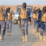 ISIS Second-In-Command Down – What’s The Truth? Camouflaged soldiers wearing masks and vests walk in desert.
