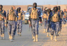 ISIS Second-In-Command Down – What’s The Truth? Camouflaged soldiers wearing masks and vests walk in desert.