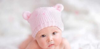 A cute baby wearing a pink knitted hat and mittens, lying on a soft surface