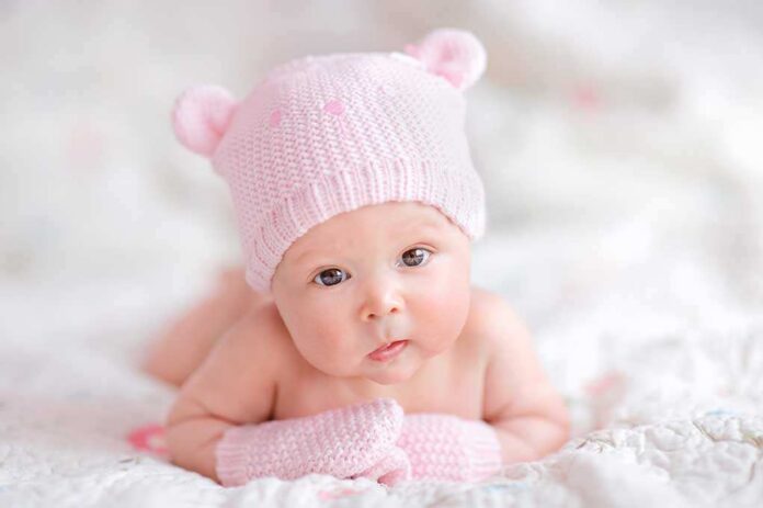 A cute baby wearing a pink knitted hat and mittens, lying on a soft surface