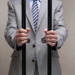 Businessman in a suit holding prison bars with handcuffs on his wrists