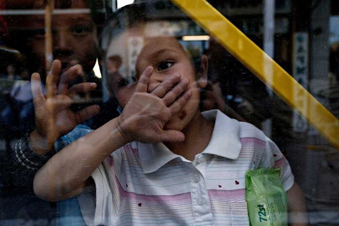 Migrants,And,Refugees,Wait,To,Be,Transported,To,The,New A child playfully covering their face while looking through a glass window