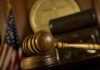 Irony Stings: Trump Critic Caves A wooden gavel resting on a desk in a courtroom with an American flag in the background