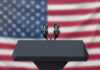 Ballot Deadline Panic Engulfs Democrats Podium with microphones, American flag background.