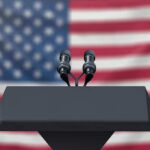 Ballot Deadline Panic Engulfs Democrats Podium with microphones, American flag background.