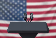 Ballot Deadline Panic Engulfs Democrats Podium with microphones, American flag background.