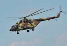 Black Hawks Swarm Camp—Why? Camouflage military helicopter in flight against blue sky.