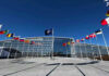 NATO Showdown: Trump Demands Greenland Flags outside NATO headquarters building under clear blue sky.