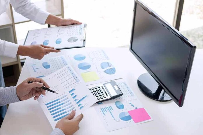 shutterstock_1221962593 Business hands reviewing charts with a calculator and monitor on desk