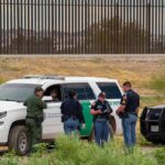 Fake Troops Run Border—Officials Stunned law enforcement officers confer beside patrol vehicles near a border fence