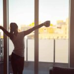 Woman stretching with coffee by a balcony window at sunrise
