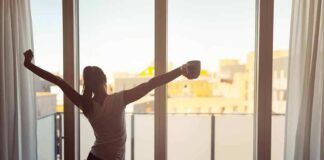 Woman stretching with coffee by a balcony window at sunrise