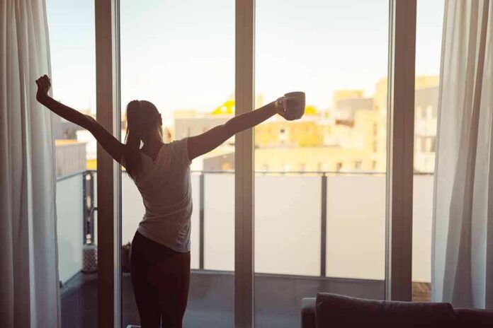 shutterstock_1016532013 Woman stretching with coffee by a balcony window at sunrise