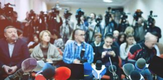 Row of microphones at a press conference with audience and cameras behind