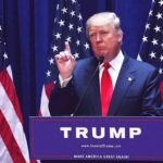man in suit speaking at podium with multiple American flags